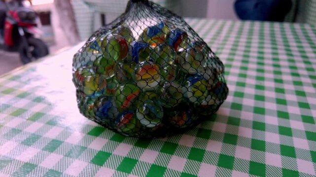 Camera slowly pulls back from a mesh bag filled with colorful glass marbles placed on a green checkered table.