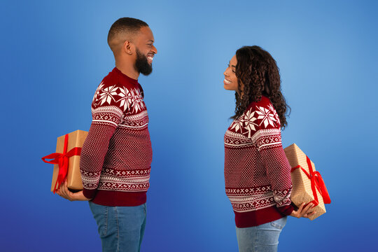Loving african american man and woman holding Christmas presents behind their backs, exchanging gifts over blue studio background. Happy black couple celebrating New Year together