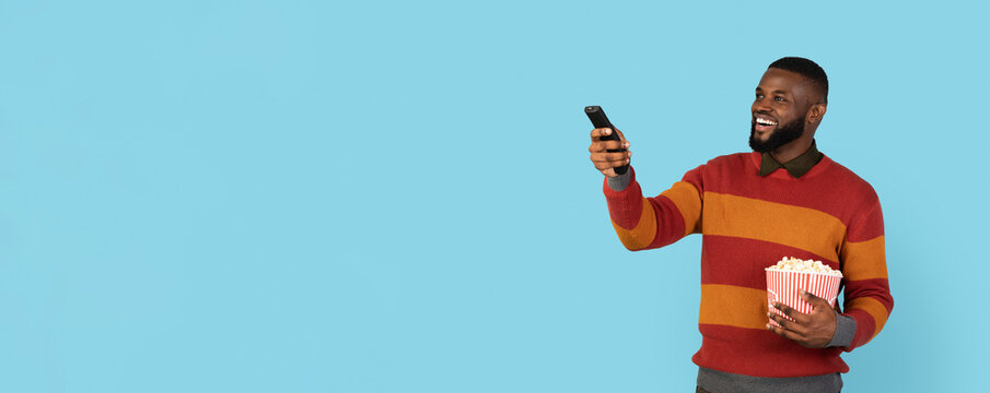 Excited young black man watching TV and eating popcorn, smiling african american man switching channels with remote controller in hand while standing isolated on blue background, panorama, copy space
