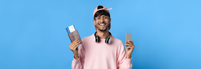 A cheerful young man stands against a bright blue background. He holds a portable charger in one hand and a smartphone in the other.