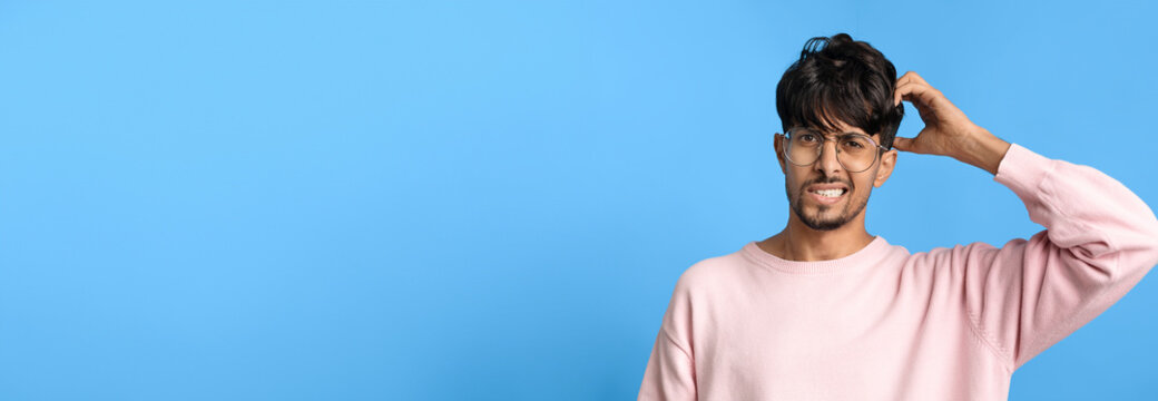 A young man appears confused as he scratches his head, wearing a light pink sweater and glasses. He stands in front of a bright blue background, expressing a puzzled emotion.
