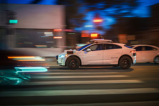 LOS ANGELES, CA - March 3, 2025: Driverless Waymo self driving taxi car driving in night city traffic, crossing illuminated street intersection at dusk in West Hollywood, California.