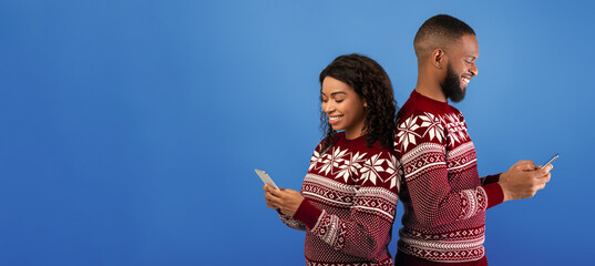 People and technology concept. Happy black couple holding smartphones and standing back to back, surfing internet on blue background, studio shot. Social network addiction