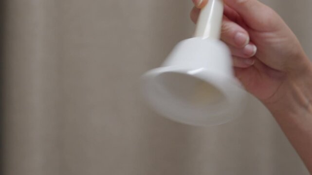 A hand rings a white, metal handbell with a white handle. The bell produces a clear, high-pitched ringing sound.