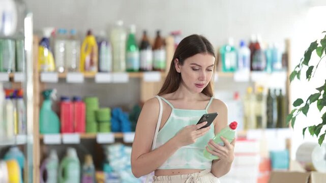 Woman scans QR code on label of detergent using her smartphone, while in the background shoppers browse products. High quality 4k footage