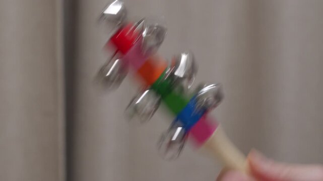 A hand shakes a colorful wooden stick with multiple metal jingle bells. The sleigh bells produce a bright, rhythmic jingling sound.