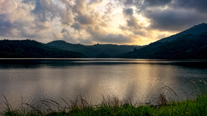 A view of a lake at the foot of the mountains in Thailand during sunset. The sun's rays filter through the clouds, creating golden reflections on the water. An atmosphere of peace and harmony.