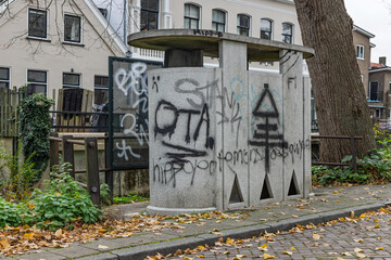 Concrete public urinal in Zutphen, Netherlands, heavily covered in graffiti, situated on a street with historical buildings in the background.