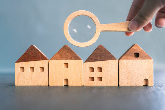 Hand holding magnifying glass over wooden houses symbolizing real estate search, property inspection, home selection, housing investment, or mortgage decision-making process.