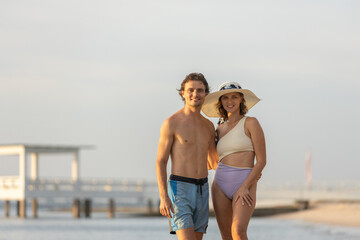 Happy couple posing on beach during summer vacation, smiling together in swimwear and sun hat. Romantic travel moment showing love, relaxation and outdoor lifestyle by the sea.