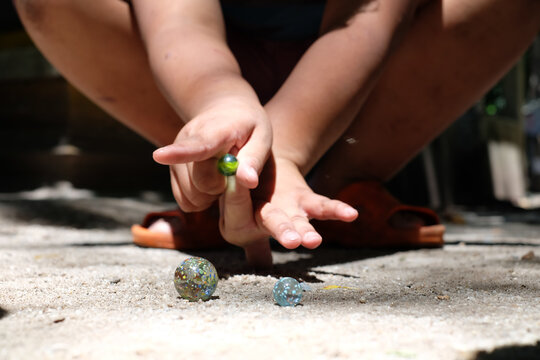 Child playing with marbles on sandy ground, focusing on aiming and shooting with concentration. scene captures playful and nostalgic moment in natural light
