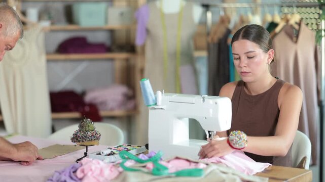 Working day in sewing workshop, textile industry. Girl master sew clothes in sewing machine, mature man pins paper pattern to fabric, attaches template to fabric, preparing to cut fabric into pieces.