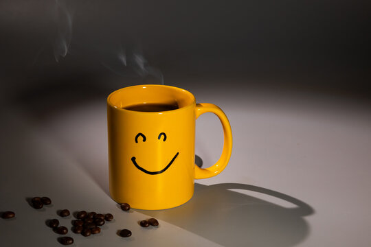 A yellow coffee mug featuring a smiley face sits on a surface, near a scattering of coffee beans. Steam rises from the mug, suggesting a warm and inviting beverage.