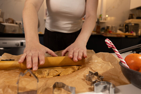 A woman in her kitchen prepares gingerbread cookies. She is rolling out the dough with a wooden rolling pin. Cookie cutters are visible on the table, plus tangerines and a candy cane.