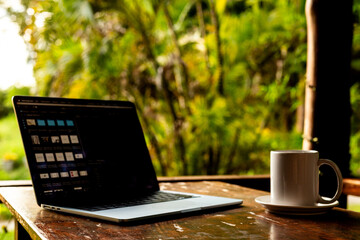 Laptop and cup on wooden table of terrace with tropical forest view at sunset, remote work digital nomad.
