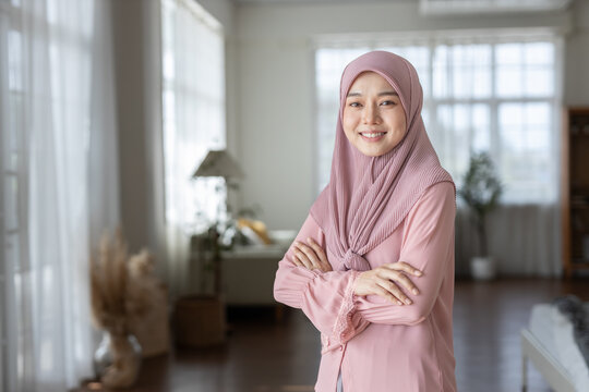 Portrait of confident Muslim woman wearing pink hijab and modest outfit, arms crossed and smiling in cozy home interior. Ideal for content about leadership, empowerment, and lifestyle diversity. - Powered by Adobe