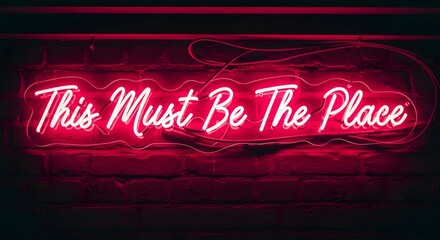A vibrant neon sign displaying the phrase 'This Must Be The Place' in bright pink color against a dark brick wall background