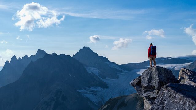 Hiker Stands on a Cliff Overlooking Jagged Mountain Range Under Blue Sky During Sunrise Adventure