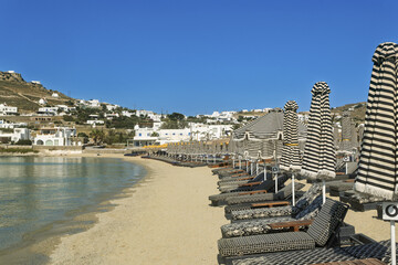 Serene beach scene in Mykonos, Greece,  with a row of black and white striped lounge chairs and matching umbrellas arranged neatly on sandy shore,