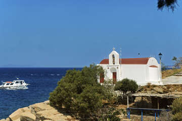 Scenic coastal view in Mykonos featuring a small white chapel with a red roof and cross on top, located near a rocky shoreline, with arched windows and a boat cruising 