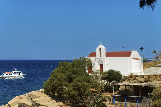 Scenic coastal view in Mykonos featuring a small white chapel with a red roof and cross on top, located near a rocky shoreline, with arched windows and a boat cruising  - Powered by Adobe