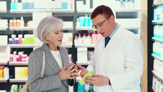 Interested old lady asking middle-aged male apothecary for advice concerning preparation in box in drugstore
