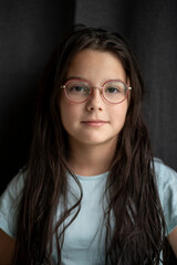 Fototapeta premium Vertical portrait of a girl with long hair and glasses, serious face against a dark fabric background, soft light, copy space. Concept of focus, depth, and natural emotion.