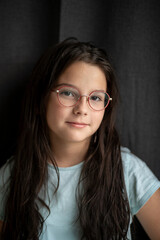 Fototapeta premium Girl with long hair and glasses slightly tilting her head, gentle half-smile, dark fabric background, soft light, copy space. Concept of emotion and natural beauty.
