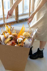 Woman in a beige dress and black boots holding a vibrant autumn flower bouquet in a large brown box near a sunny window indoors