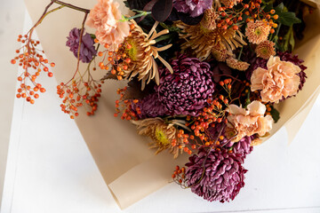 Lush autumn floral bouquet featuring purple chrysanthemums, peach carnations, and vibrant orange rowan berries wrapped in natural kraft paper on a white surface