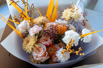 Autumnal floral arrangement featuring peach roses, yellow chrysanthemums, protea, and orange accents, neatly packaged in a brown craft box by a window
