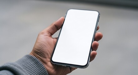 A hand holding a smartphone with a blank white screen against a blurred neutral background outdoors