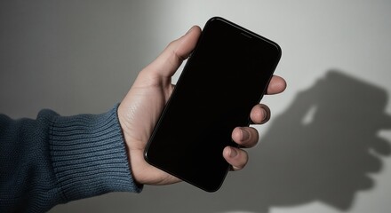 A hand holding a black smartphone with a blank screen against a neutral background with a shadow casted