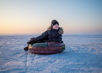 Obraz premium young boy in winter attire rests on an inflatable snow tube in a vast, snow-covered landscape under a soft, pastel-colored sky.