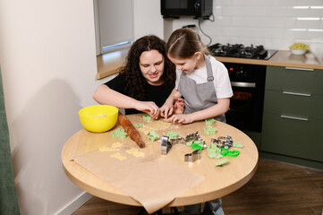 Mother and daughter bake cookies in kitchen. Woman enjoys baking with daughter and enjoying cutting out cookies from dough with cookie cutters in kitchen. Cooking at home, childhood hobby.