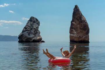 Woman, sea, rocks: A happy woman floating on an inflatable ring in the clear sea with two large rock formations and blue sky.