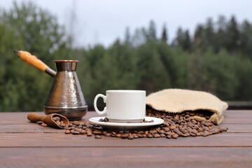 Against the backdrop of the forest, on a brown wooden table there is a white cup of coffee, next to it a copper cezve, a scattering of coffee beans from burlap sack and wooden scoop with coffee beans.