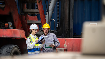Two workers are standing next to a red truck, looking at a tablet. They are wearing safety gear and seem to be discussing something important