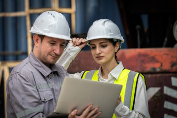 Two people wearing hard hats are looking at a laptop. One of them is a woman. They are looking at a document on the laptop