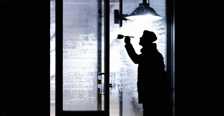 Fototapeta premium Silhouette person brushes snow from glass door. Winter scene, dark exterior, bright interior light visible. Cold weather conditions present.