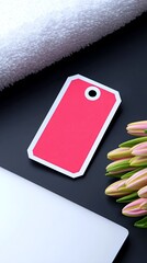 A flat lay composition featuring a pink tag, a white folded towel, a laptop, and a bouquet of pink tulips on a dark surface.