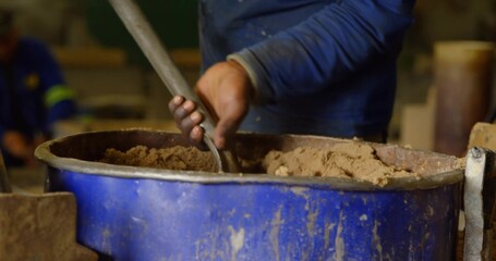 Mixing worker's hands in blue shirt pushing rod into brown mixture at workshop, with blue tub