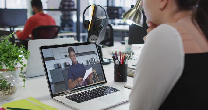Watching remote presenter wearing dark top holding papers on laptop at office desk, lamp