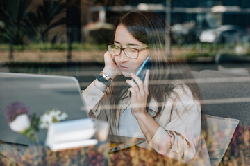 Woman with glasses on phone call working on laptop behind cafe window
