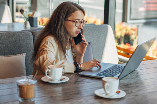 Woman on phone call working on laptop with coffee in modern cafe
