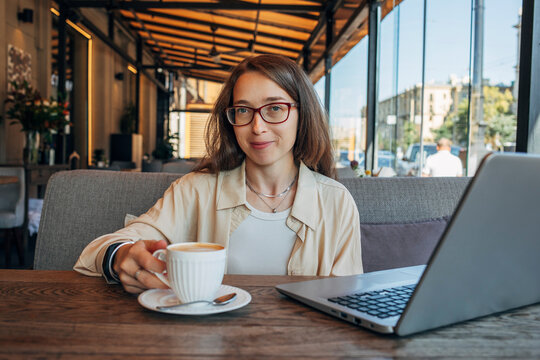 Woman with glasses holding coffee by laptop in sunlit cafe