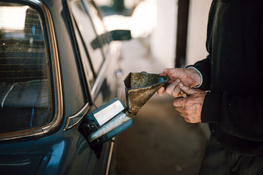 Man fills old blue car with fuel using a metal funnel