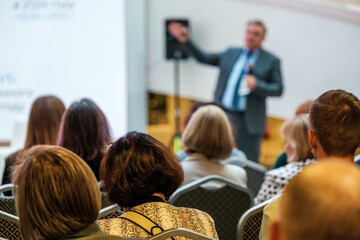 Professional conference scene with speaker in suit addressing audience in bright venue attendees seated and listening creating focused atmosphere teamwork learning vibe