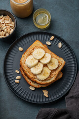 Peanut butter toast and banana on a black plate on a dark background with nuts