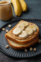 Peanut butter toast and banana on a black plate on a dark background with nuts